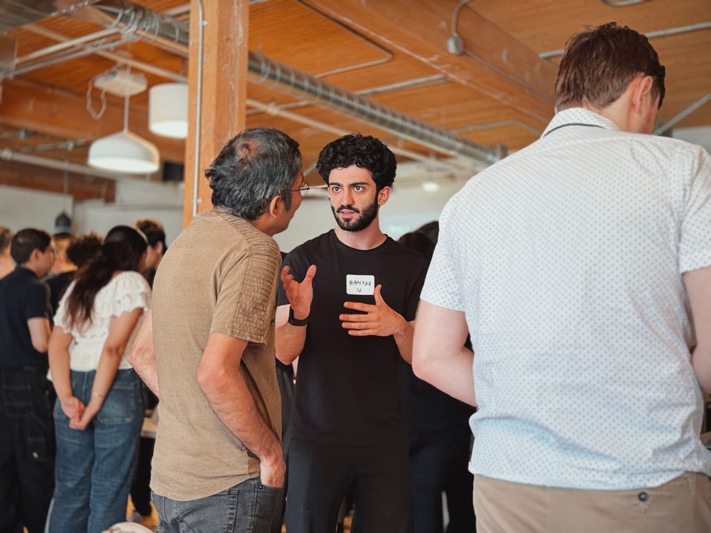 Mahyar, in a black T-shirt and a “MAHYAR 16” name tag, talking with a visitor in a crowded room.